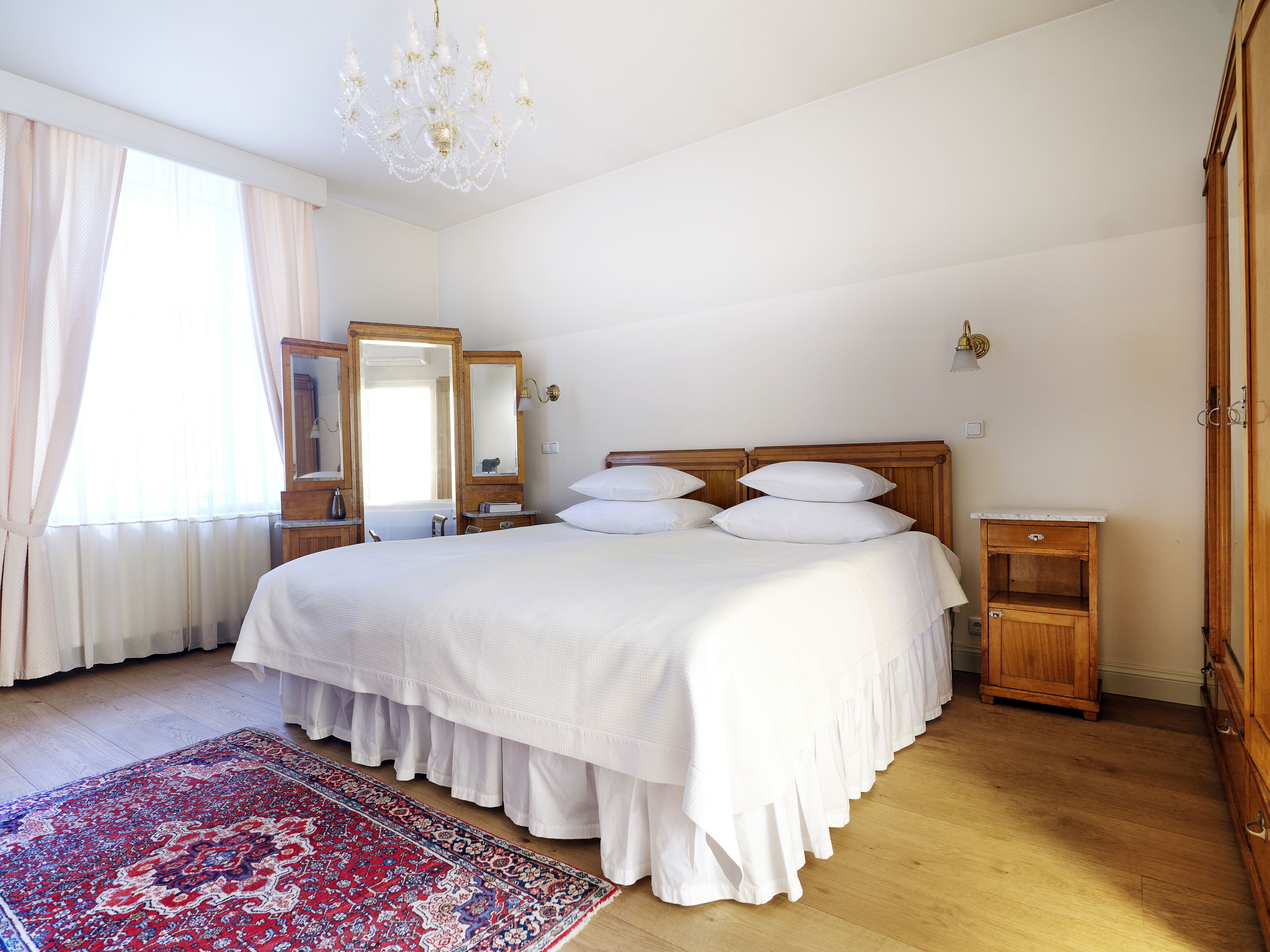 A bedroom with a large bed, a wooden nightstand, a mirror, a chandelier, and a red and white patterned rug.