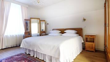 A bedroom with a large bed, a wooden nightstand, a mirror, a chandelier, and a red and white patterned rug.