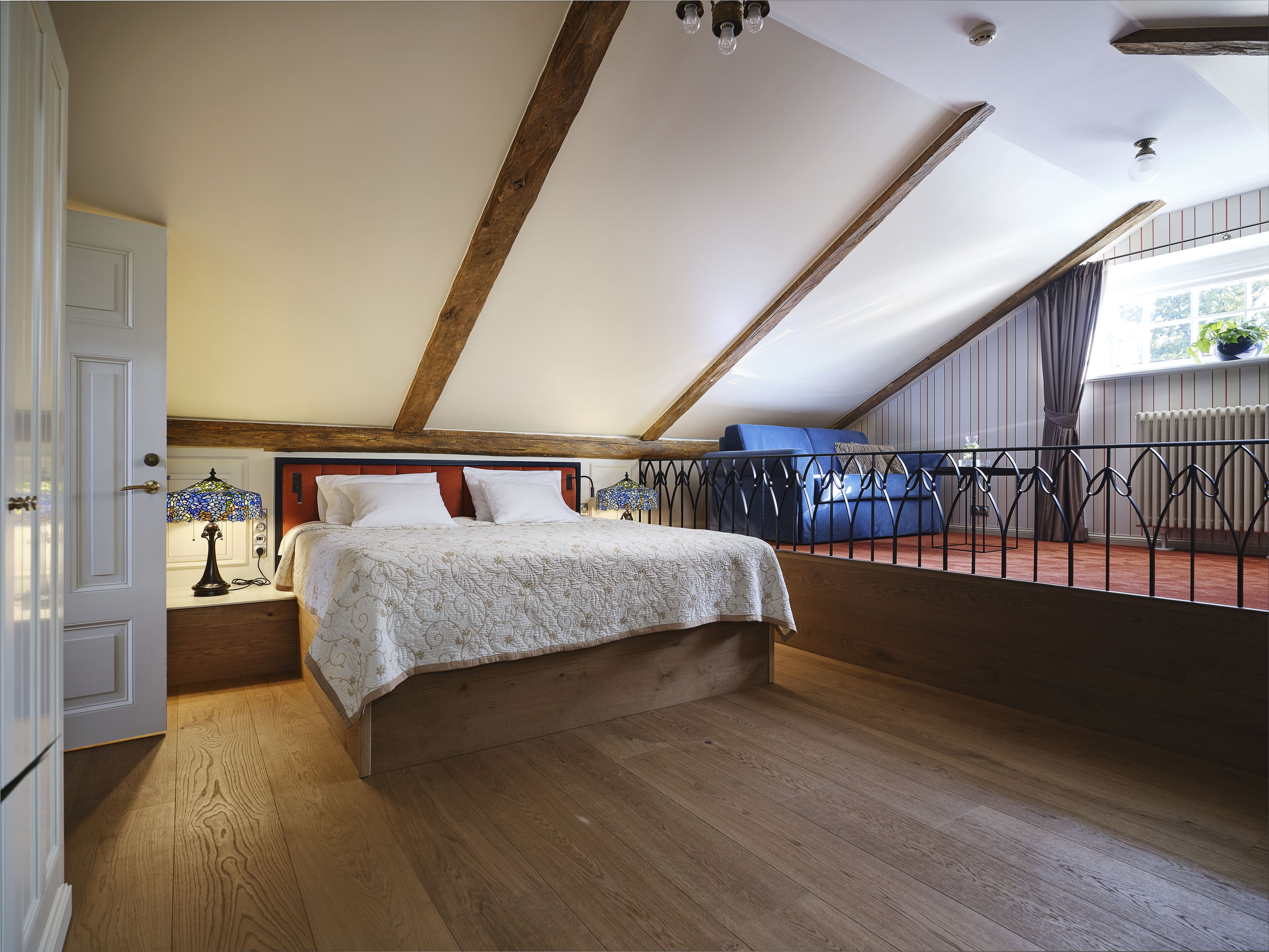 A bedroom with a wooden floor, a bed with a patterned bedspread, a nightstand with a lamp, and a view of a balcony with a railing.