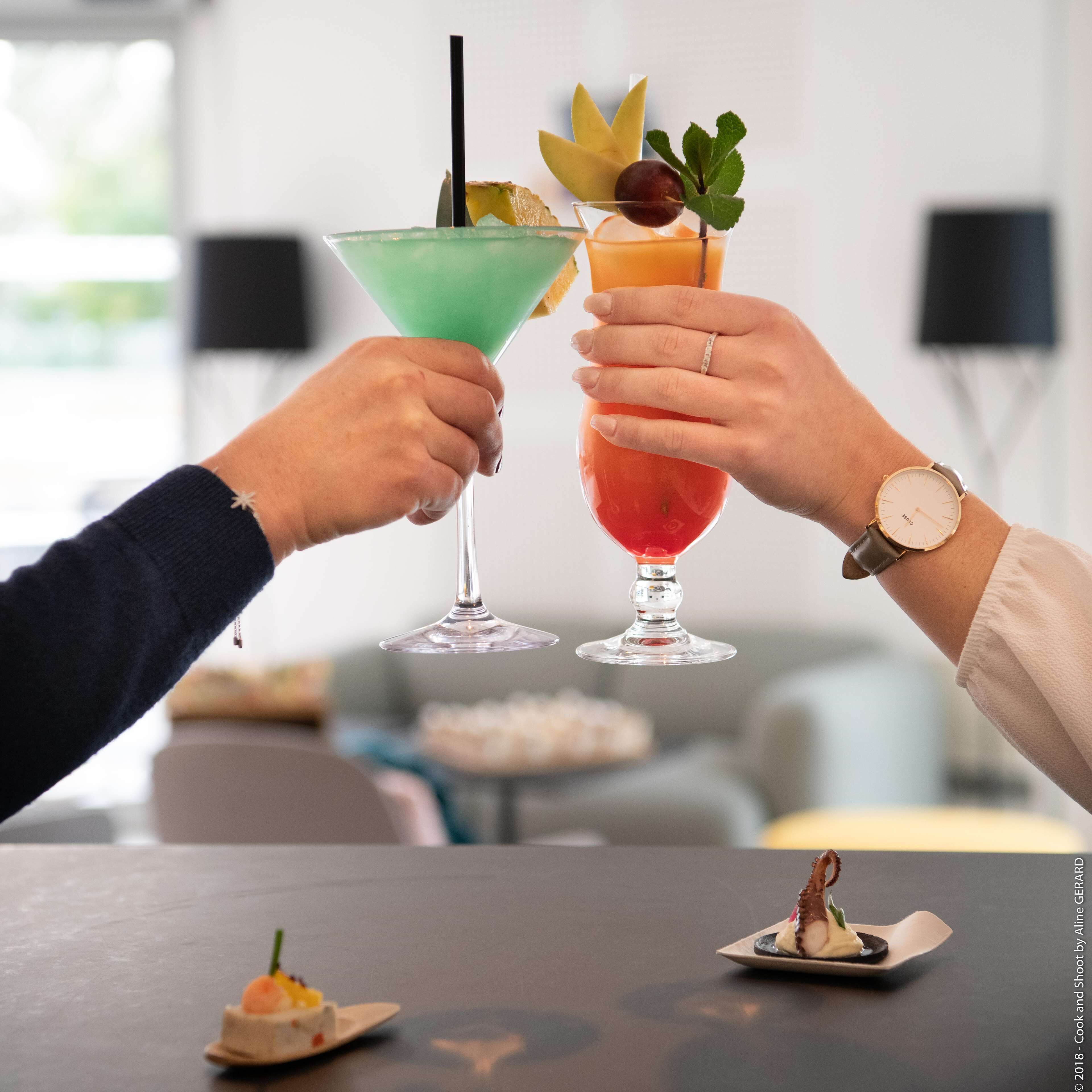Two people toasting with cocktails at a table with a dessert and a small plate of food in the background.