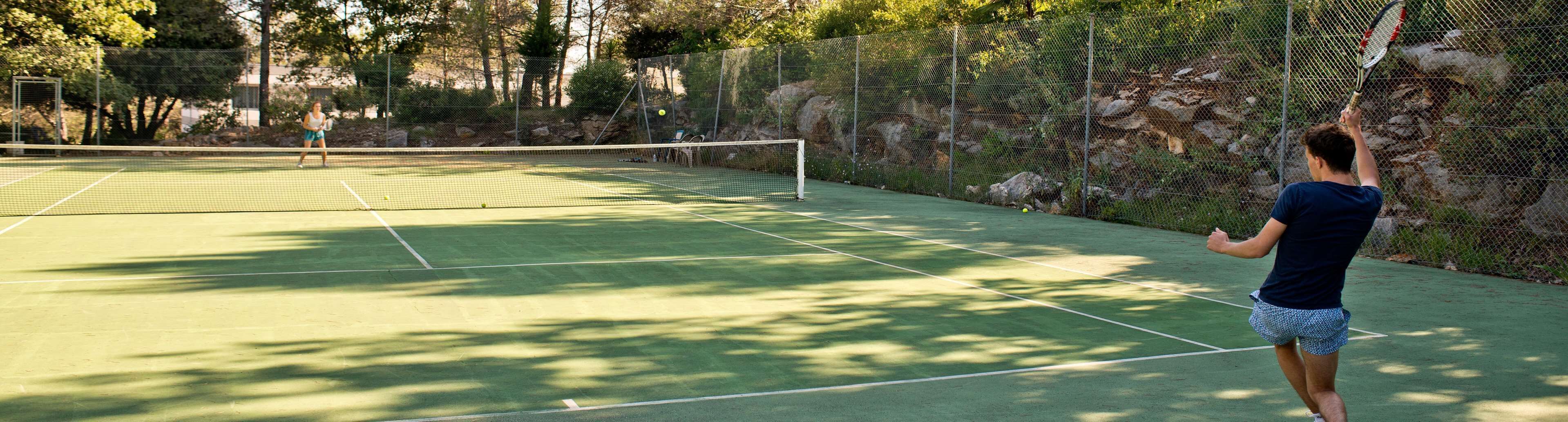 An outdoor tennis court with a net, surrounded by a chain-link fence and trees.