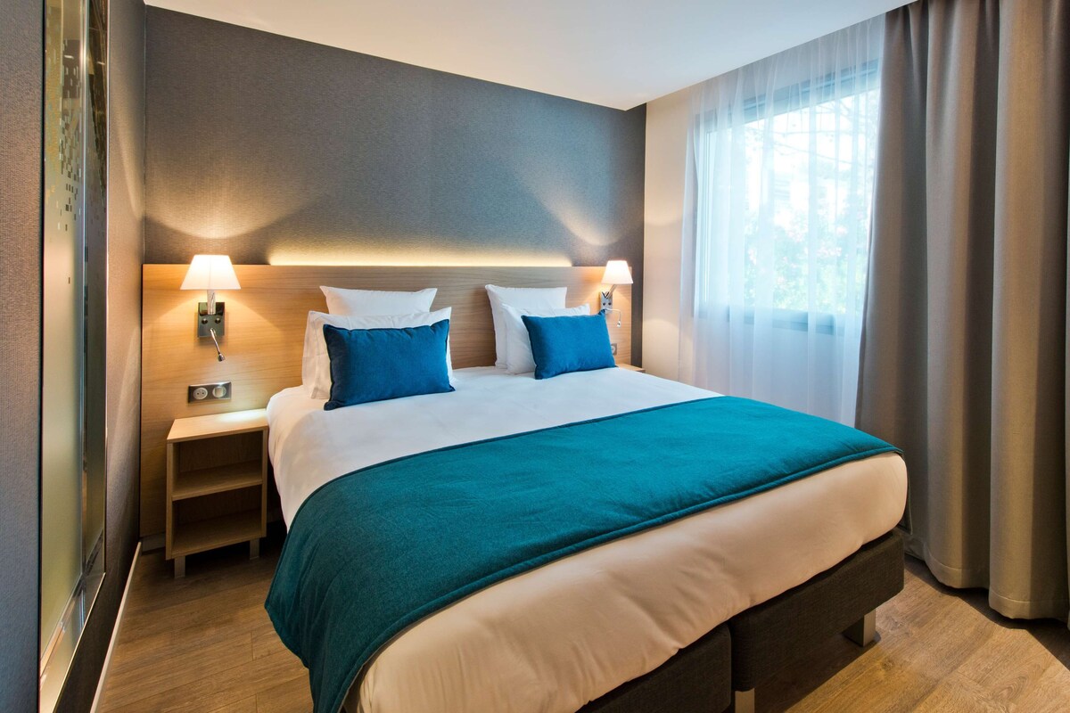 Quarto de hotel com cama, mesas de cabeceira, luminárias, janela com cortinas e vista para o verde lá fora.