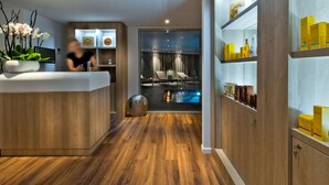 Un moderno vestíbulo de hotel con mostrador de recepción, suelo de madera, zona de piscina visible a través de una división de vidrio y estantes con artículos de aseo.
