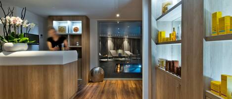 A modern hotel lobby with a reception desk, wooden flooring, a pool area visible through a glass partition, and shelves with toiletries.