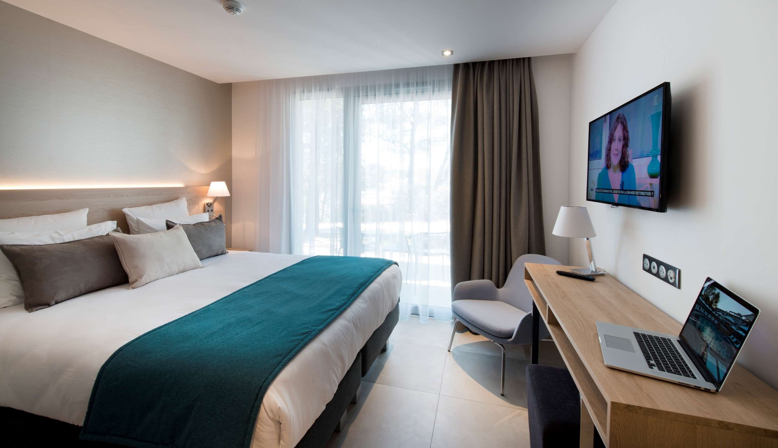 Una habitación de hotel con cama, un escritorio con una computadora portátil, un televisor y una ventana con cortinas.