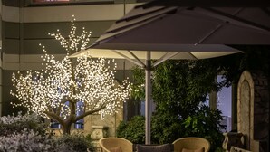 Un área de estar al aire libre con sillas de mimbre y una mesa, un árbol decorativo con luces y un muro de piedra al fondo.