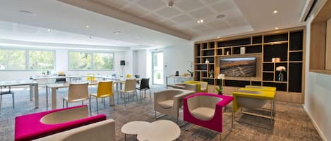 Un espacio de oficina moderno y bien iluminado, con un televisor grande, asientos cómodos y una cocina visible.