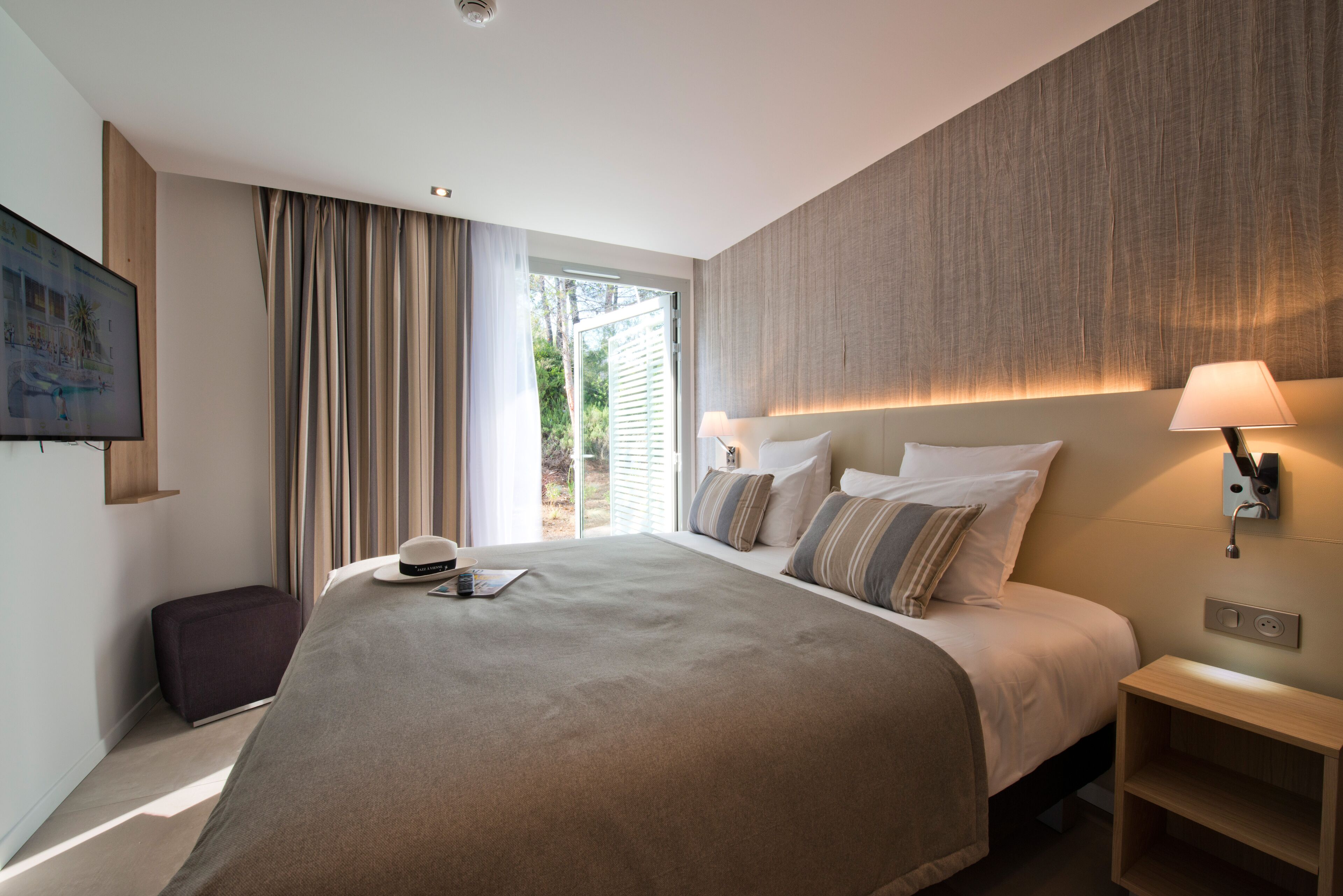 A modern hotel room with a large bed, a bedside table, a lamp, and a view of the outdoors.