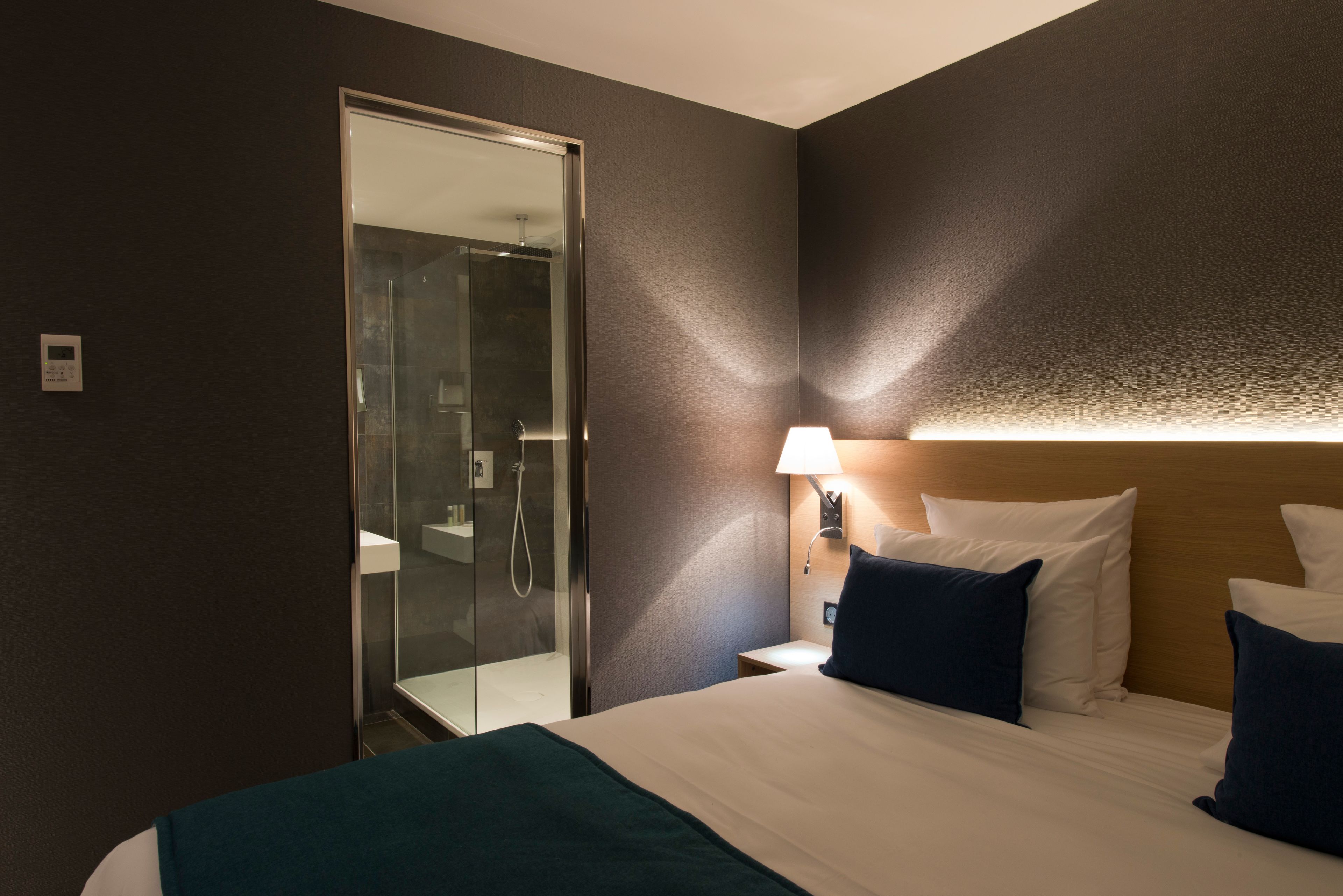 Habitación de hotel con cama, mesita de noche, lámpara y cabina de ducha de vidrio.