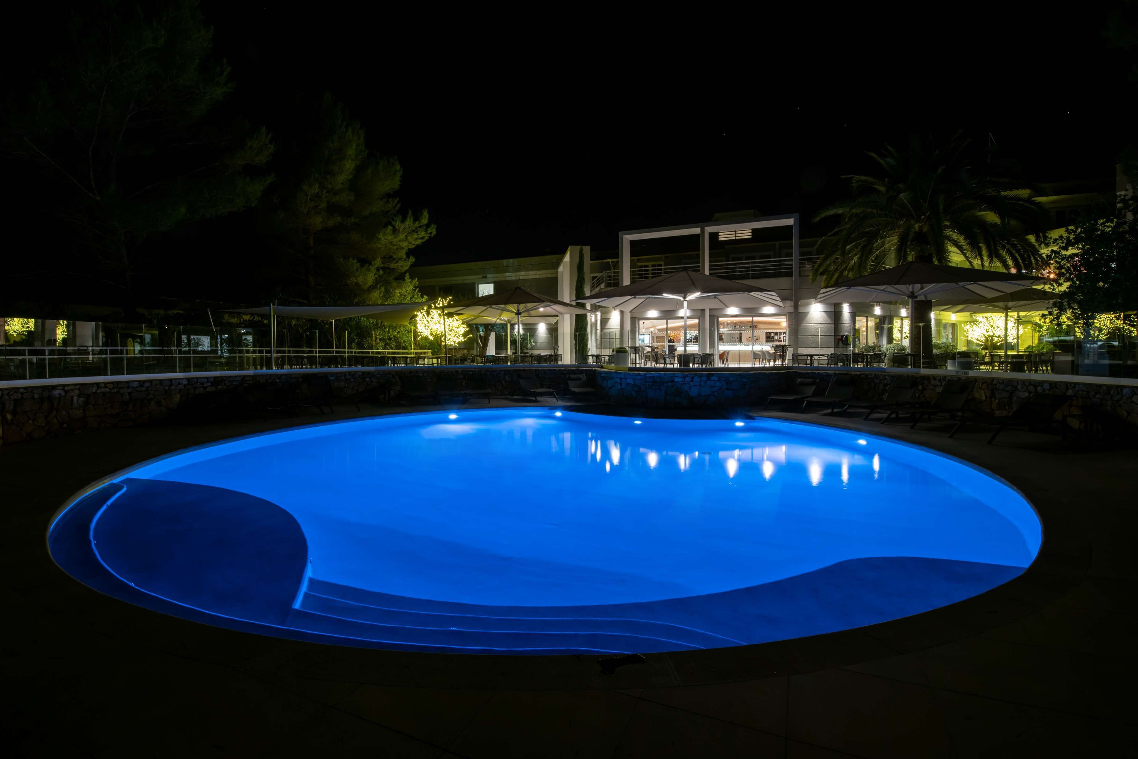 A swimming pool with a curved edge, surrounded by a stone wall and a building with outdoor seating and umbrellas in the background.