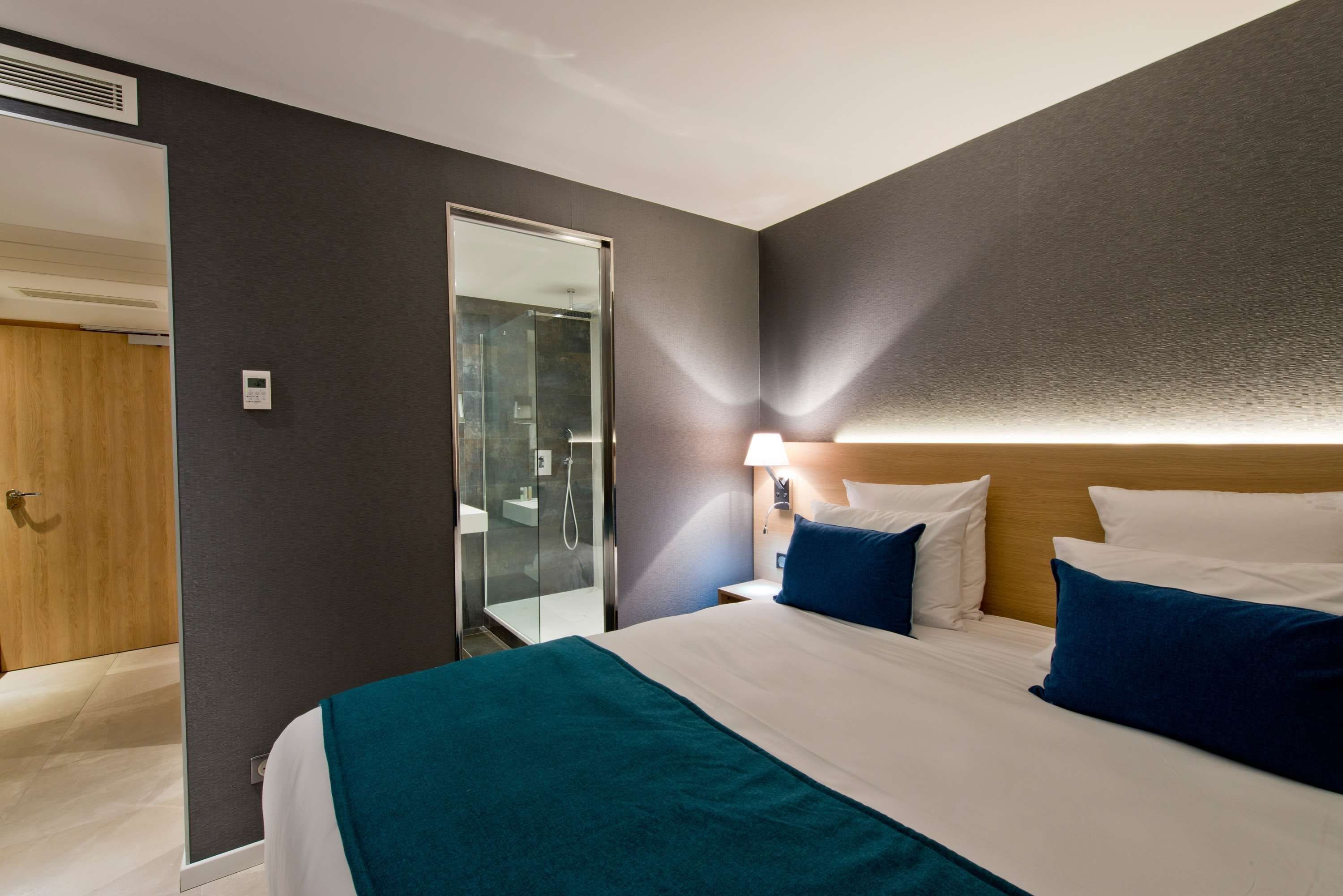 A hotel room with a bed, a nightstand, a lamp, a mirror, and a door leading to a bathroom.