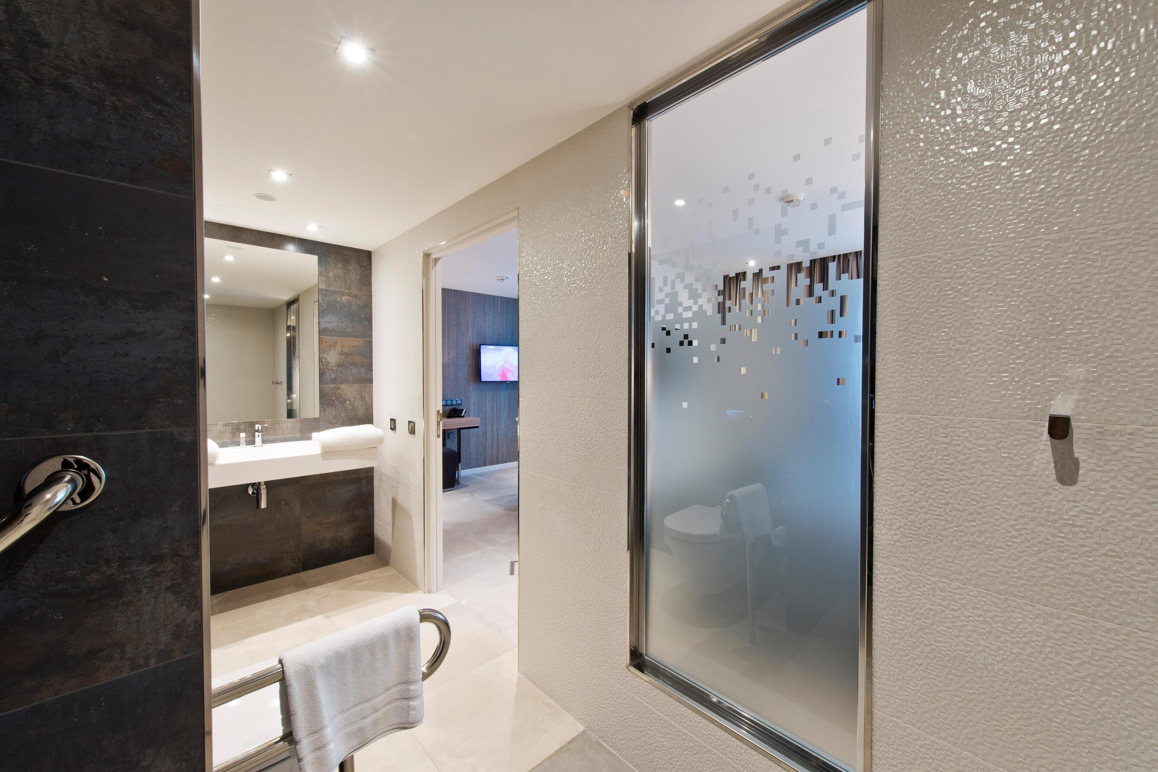 A modern bathroom with a glass shower enclosure, a separate sink area, and a visible television in the background.