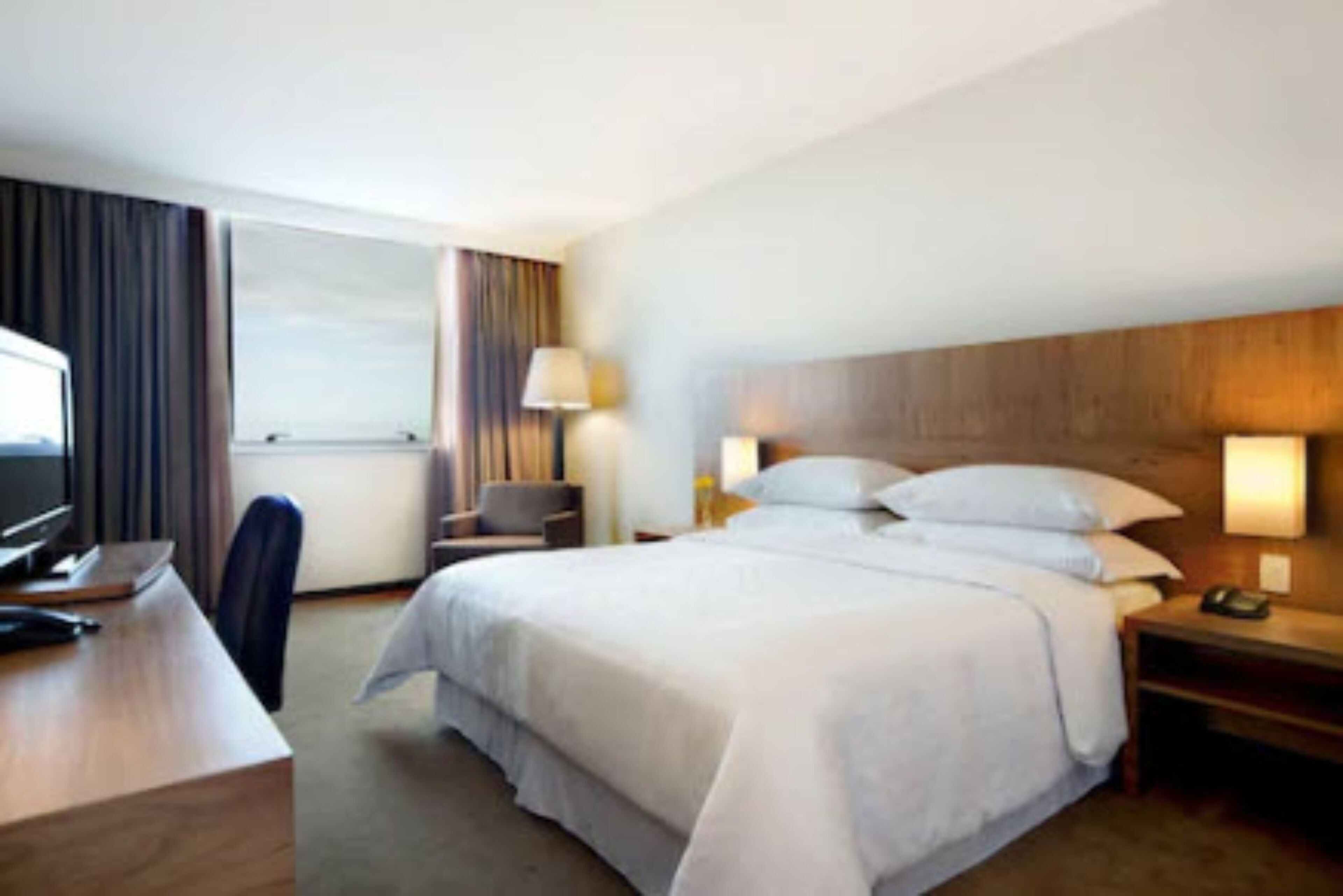 A hotel room with a large bed, a desk with a computer, a chair, and a window with curtains.