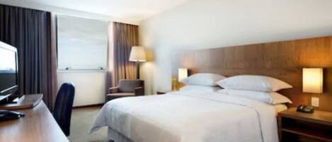 Habitación de hotel con una cama grande, un escritorio con computadora, una silla y una ventana con cortinas.