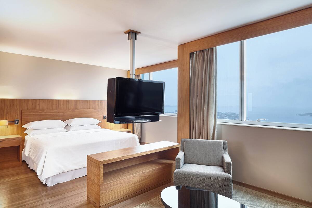 Ein Hotelzimmer mit Bett, Fernseher, Sessel und einem großen Fenster mit Ausblick.