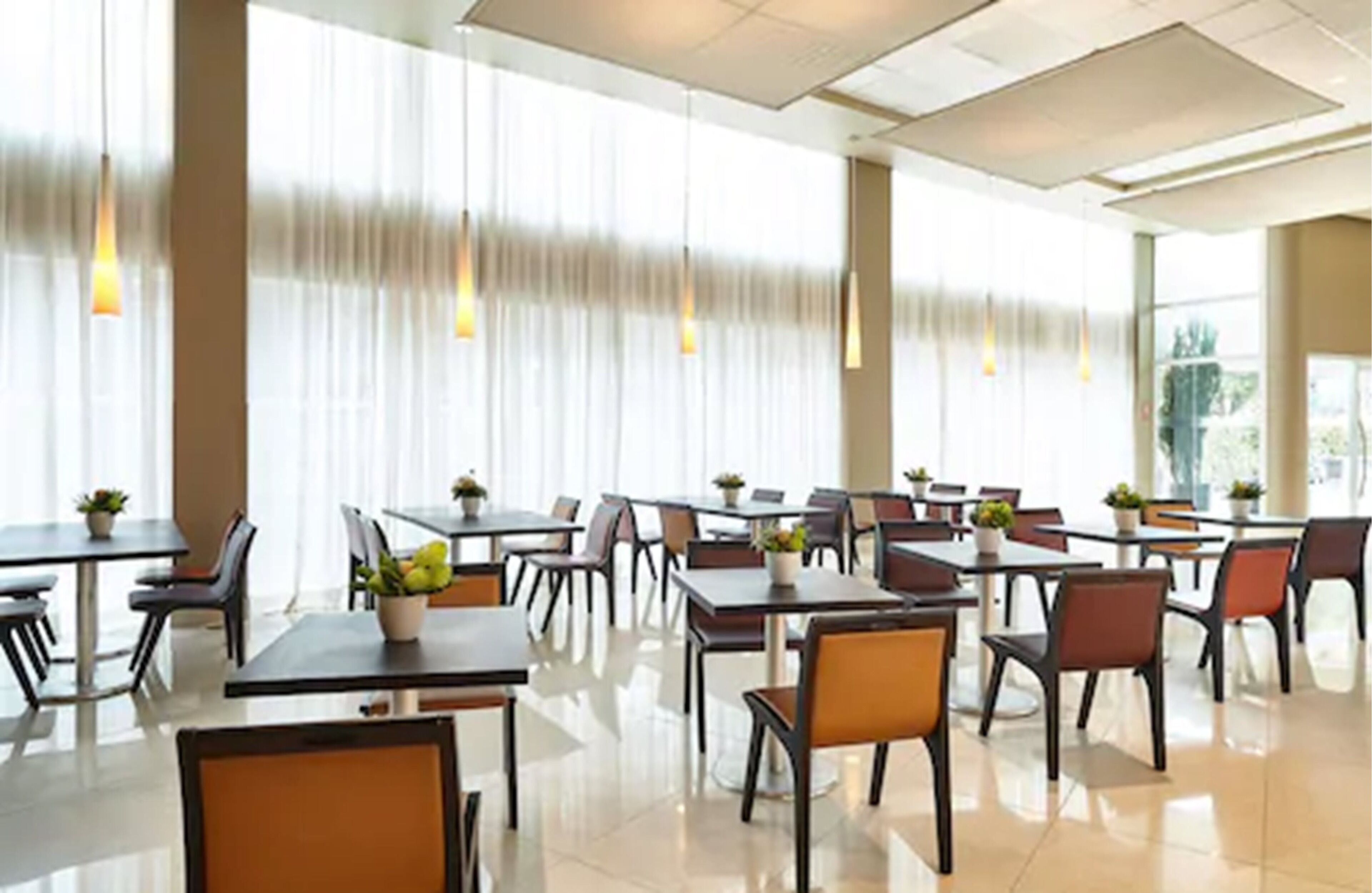 Un comedor amplio con varias mesas y sillas, ventanas grandes con cortinas translúcidas y lámparas colgantes.