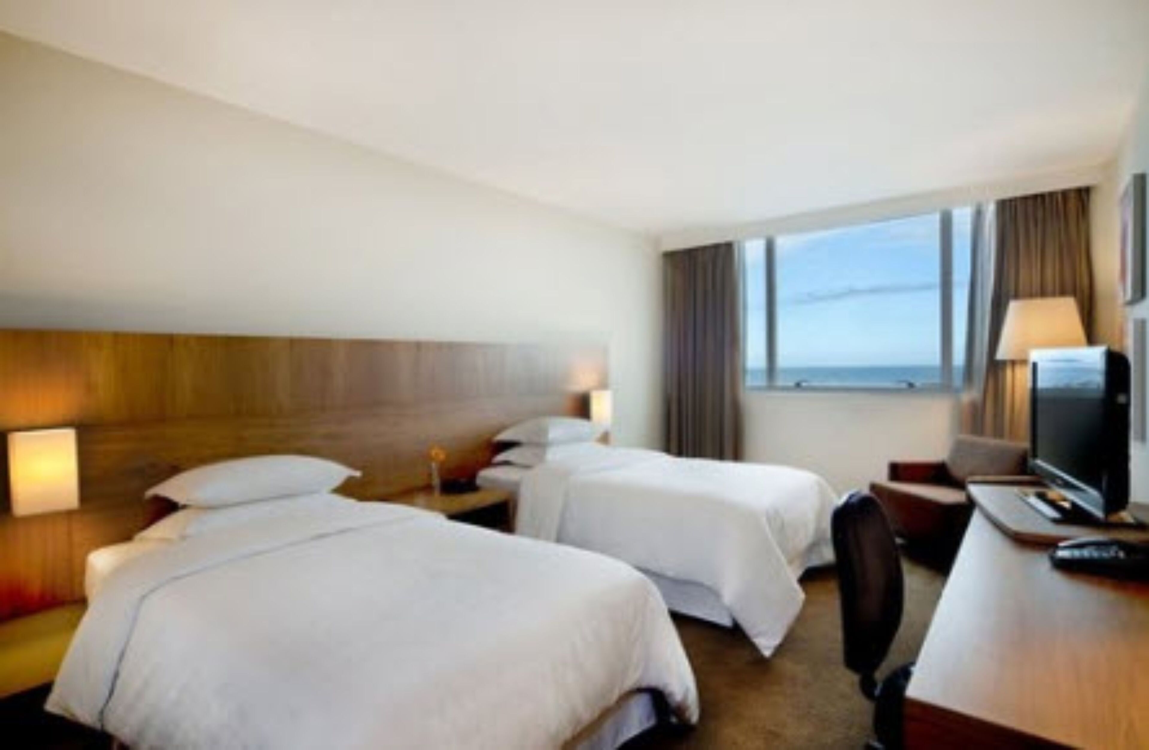 A hotel room with two beds, a desk with a television, and a view of the sea.
