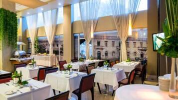 A restaurant with tables set for dining, large windows with curtains, and a view of a building outside.