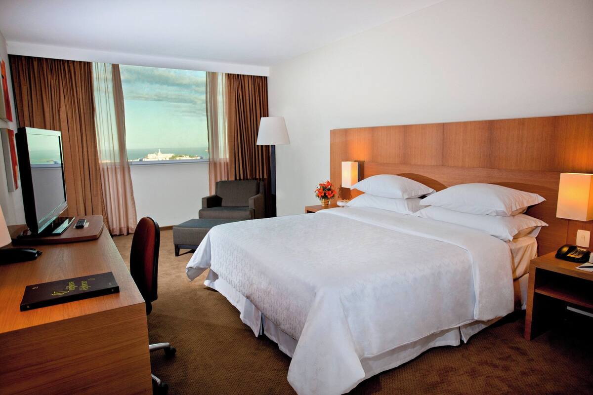 Ein Hotelzimmer mit einem großen Bett, einem Schreibtisch mit Computer, einem Sessel und Blick auf die Stadt durch das Fenster.