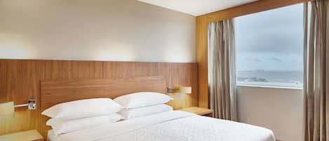 A hotel room with a wooden headboard, a large bed with white linens, a bedside table with a lamp, and a view of the sea through a window with curtains.