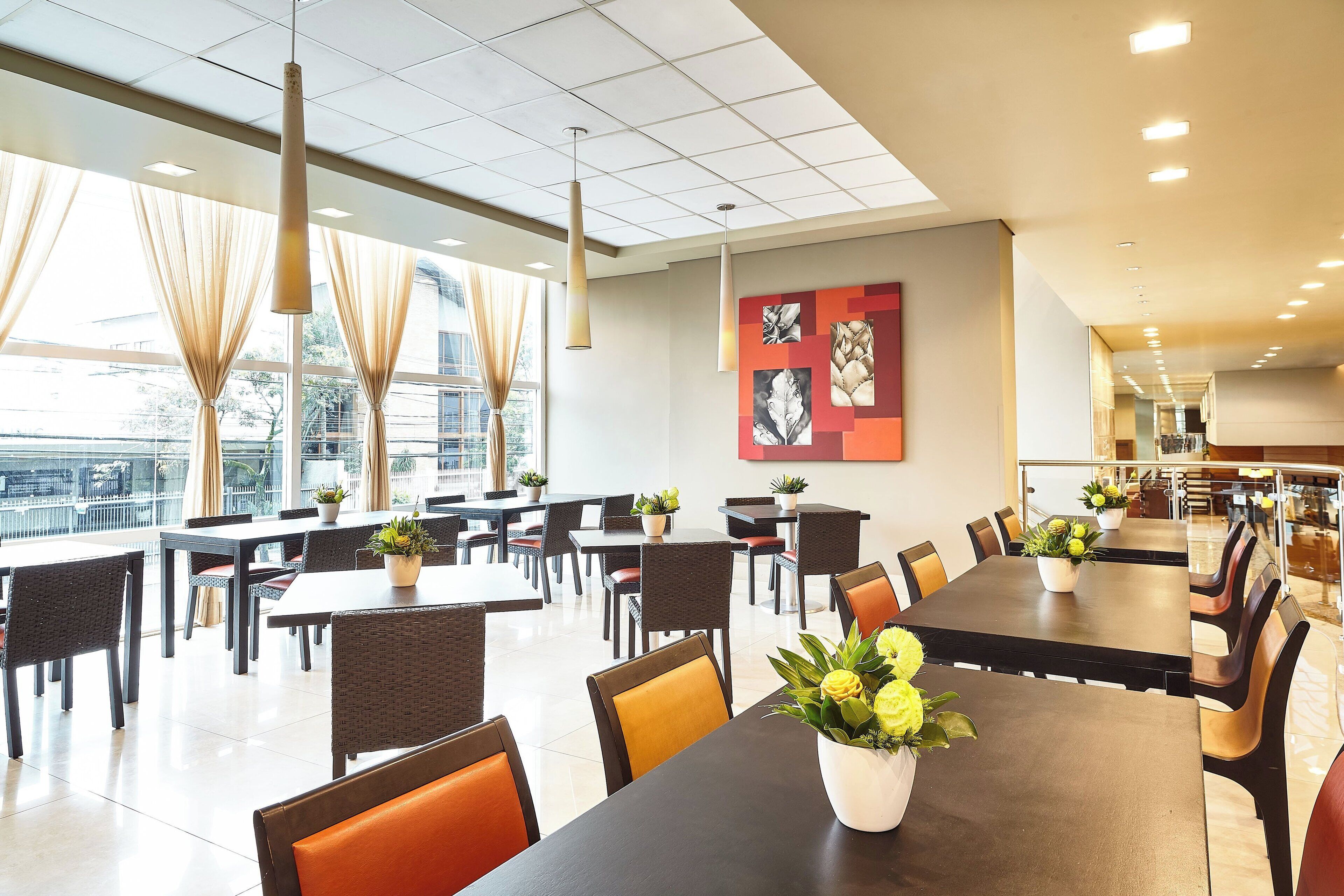 A spacious dining area with multiple tables and chairs, large windows with curtains, and ceiling lights.