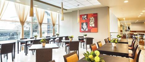 A spacious dining area with multiple tables and chairs, large windows with curtains, and ceiling lights.