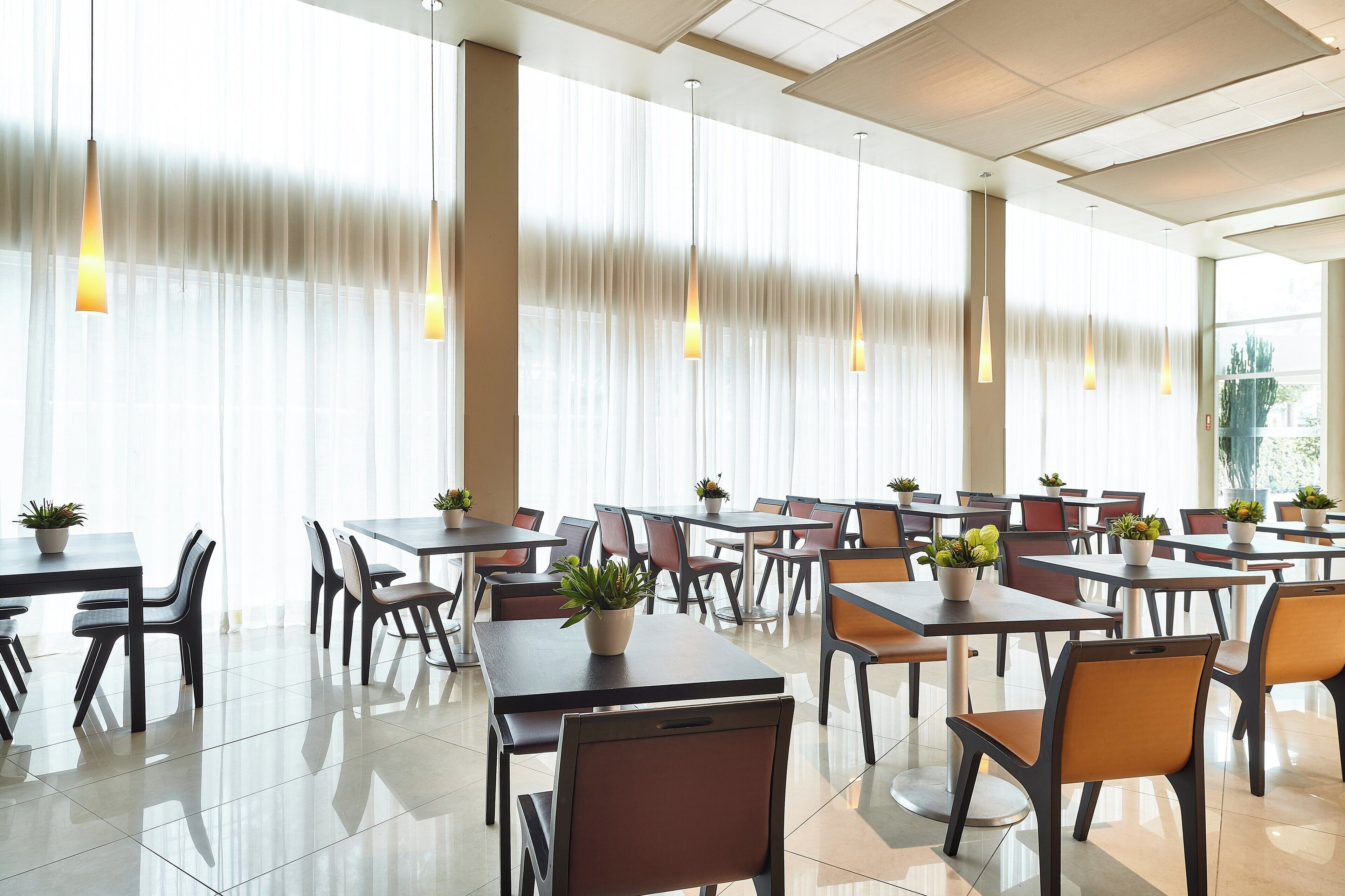 A spacious dining area with multiple tables and chairs, large windows with sheer curtains, and hanging pendant lights.