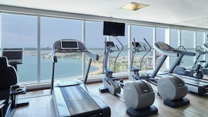 Un gimnasio con cintas de correr, elípticas y bicicletas estáticas, con vistas panorámicas al agua y al horizonte urbano.