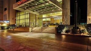 Entrada de un hotel Sheraton con fachada de vidrio, escalones que conducen a las puertas y exterior bien iluminado.
