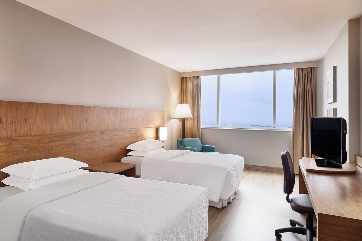 Ein Hotelzimmer mit zwei Betten, einem Schreibtisch mit Computer, einer Lampe und Blick auf die Stadt.