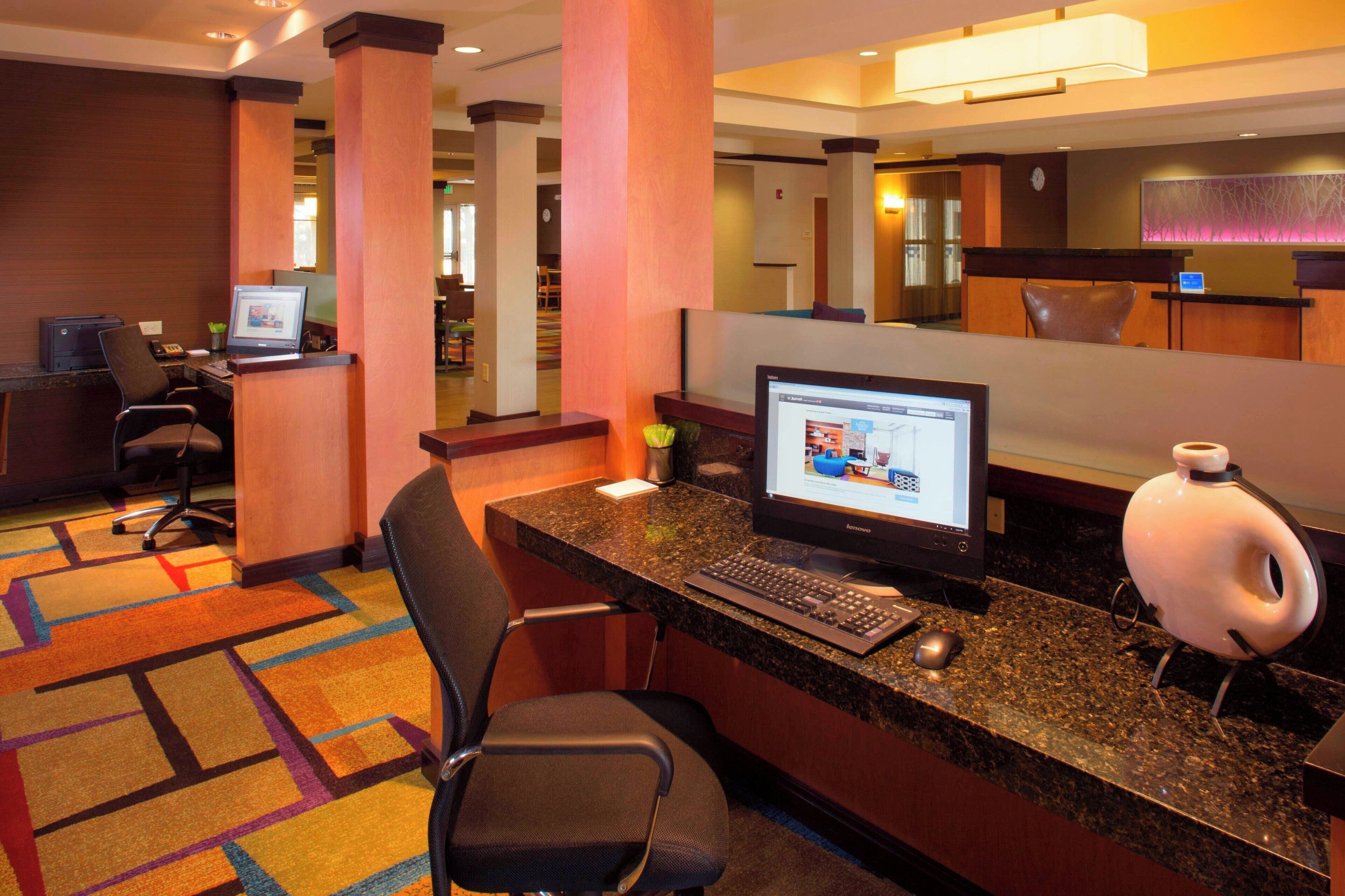 Un espace d’accueil d’hôtel comprenant un bureau avec un ordinateur, une chaise, une lampe de bureau et un sol recouvert de moquette.