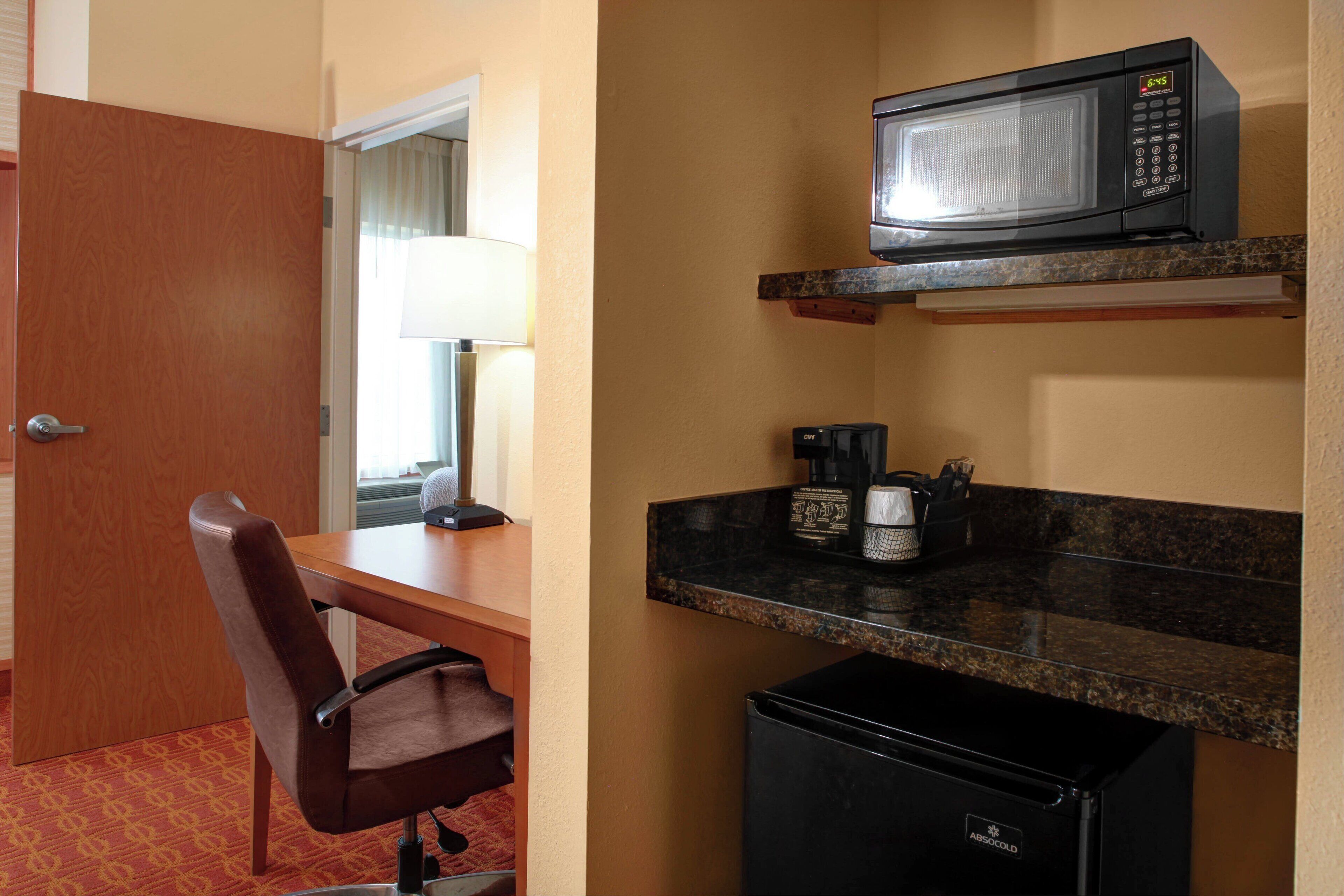 Habitación de hotel pequeña con puerta de madera, un escritorio y una silla, un microondas en la encimera, una cafetera y un refrigerador pequeño.
