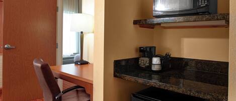 Habitación de hotel pequeña con puerta de madera, un escritorio y una silla, un microondas en la encimera, una cafetera y un refrigerador pequeño.