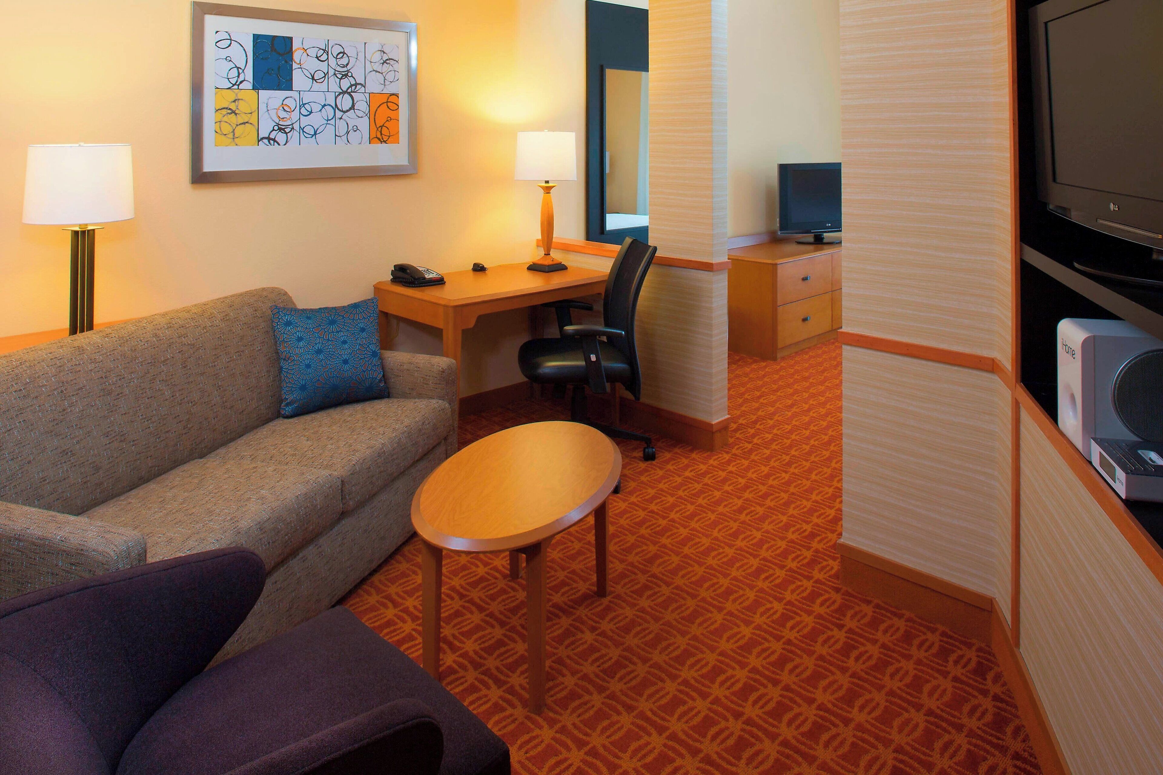 Habitación de hotel con sofá, escritorio con lámpara, silla, televisor y minibar.