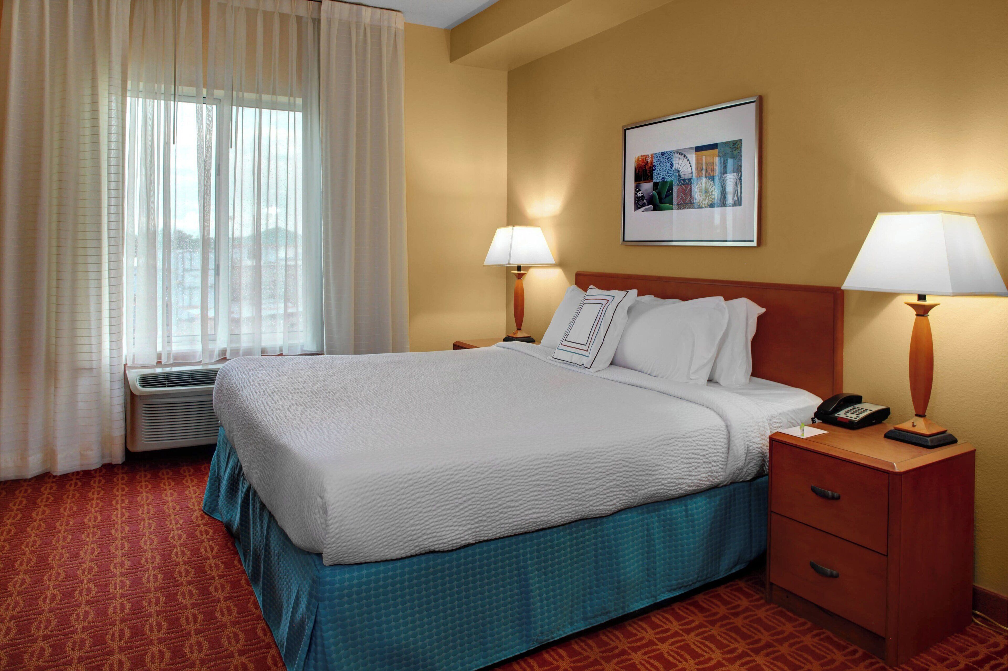 Habitación de hotel con una cama grande, dos mesitas de noche con lámparas, un cuadro en la pared y una ventana con cortinas.