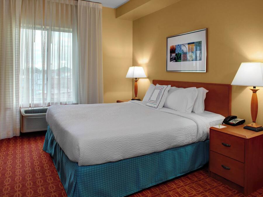 Quarto de hotel com uma cama grande, duas mesas de cabeceira com abajures, um quadro na parede e uma janela com cortinas.