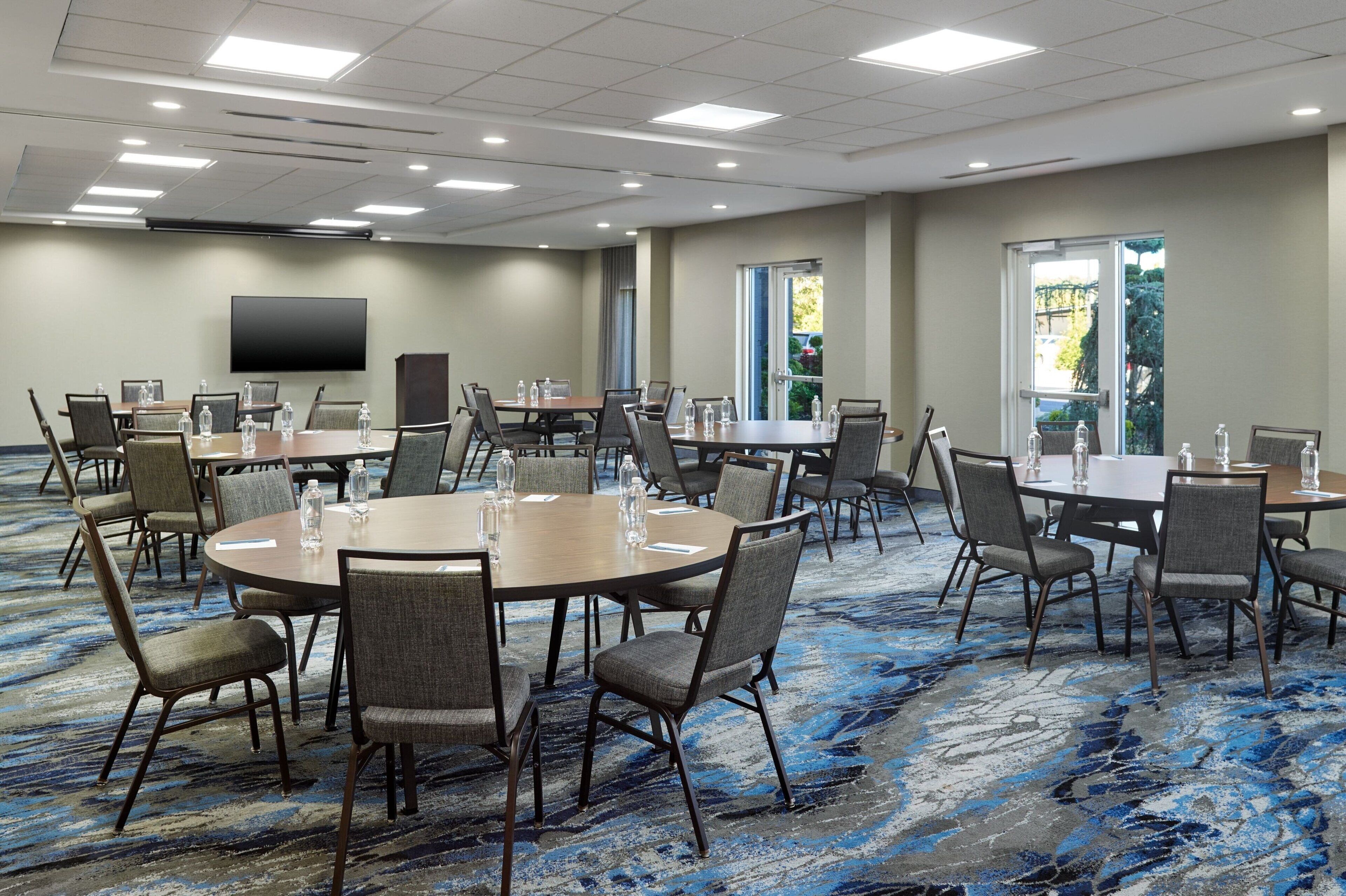A conference room with multiple tables and chairs, a flat-screen TV on the wall, and large windows with a view of greenery.