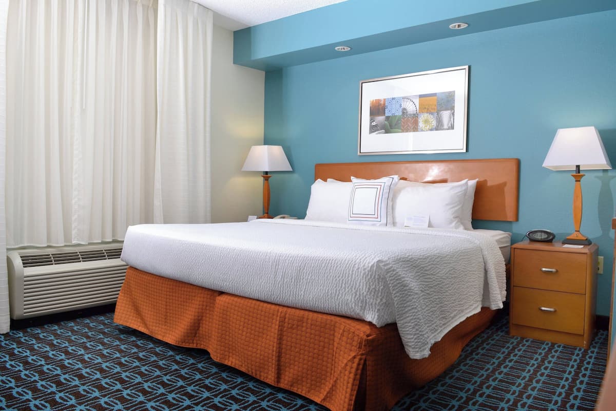 A hotel room with a bed, two bedside tables, a nightstand, a window with curtains, and a wall-mounted picture.