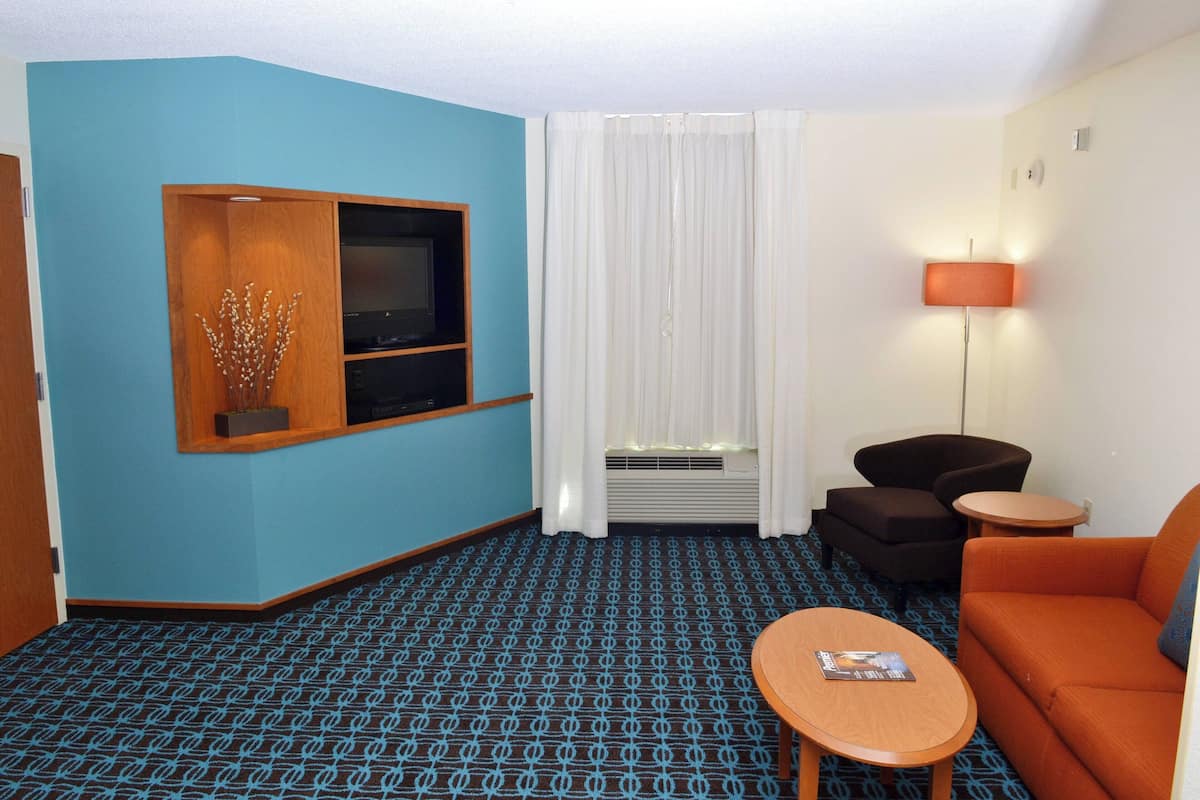 A hotel room with a blue wall, a TV on a wooden shelf, a white curtain, a lamp, a brown sofa, a small round table, and a patterned carpet.