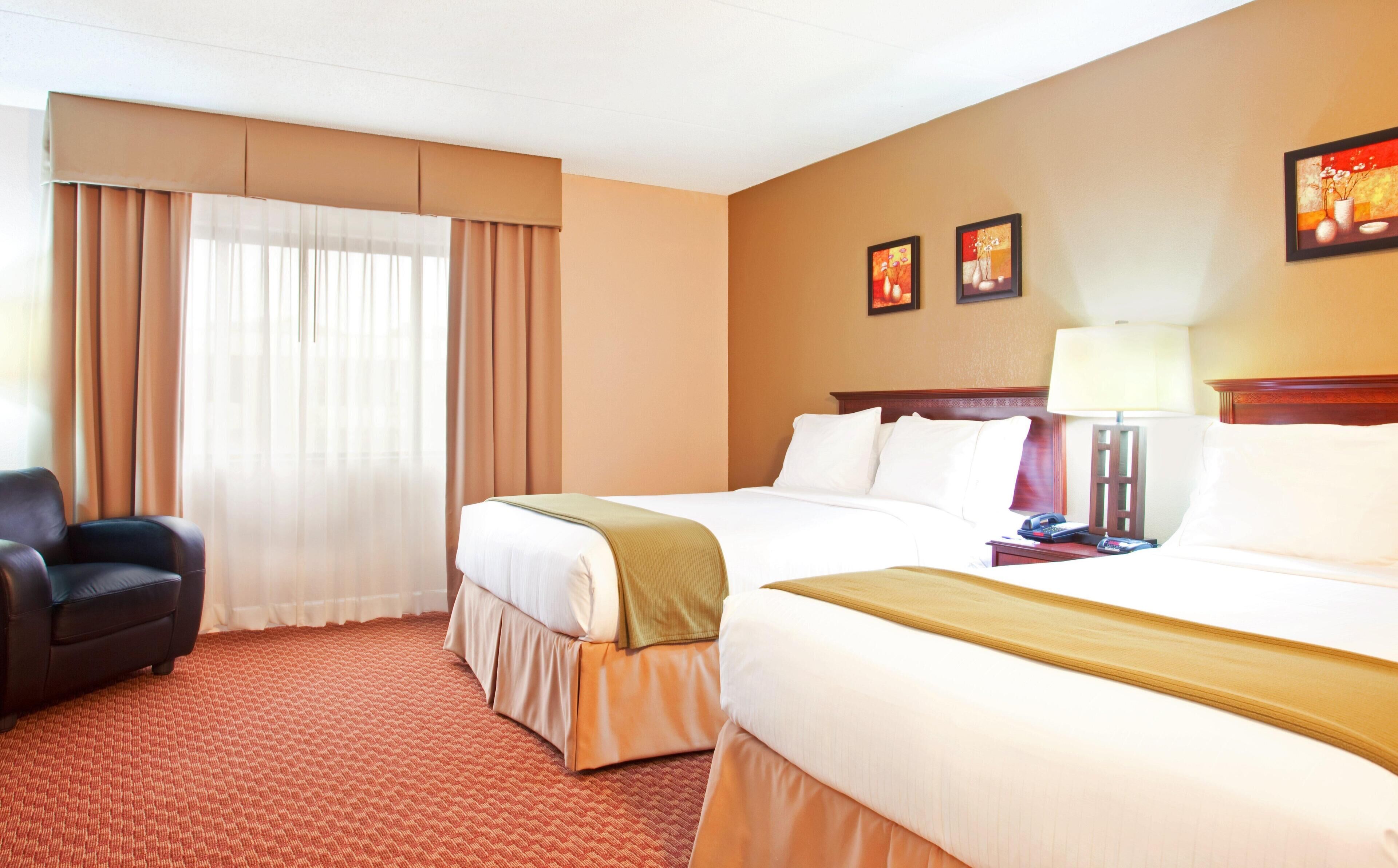 A hotel room with two beds, a chair, a desk with a lamp, and a window with curtains.