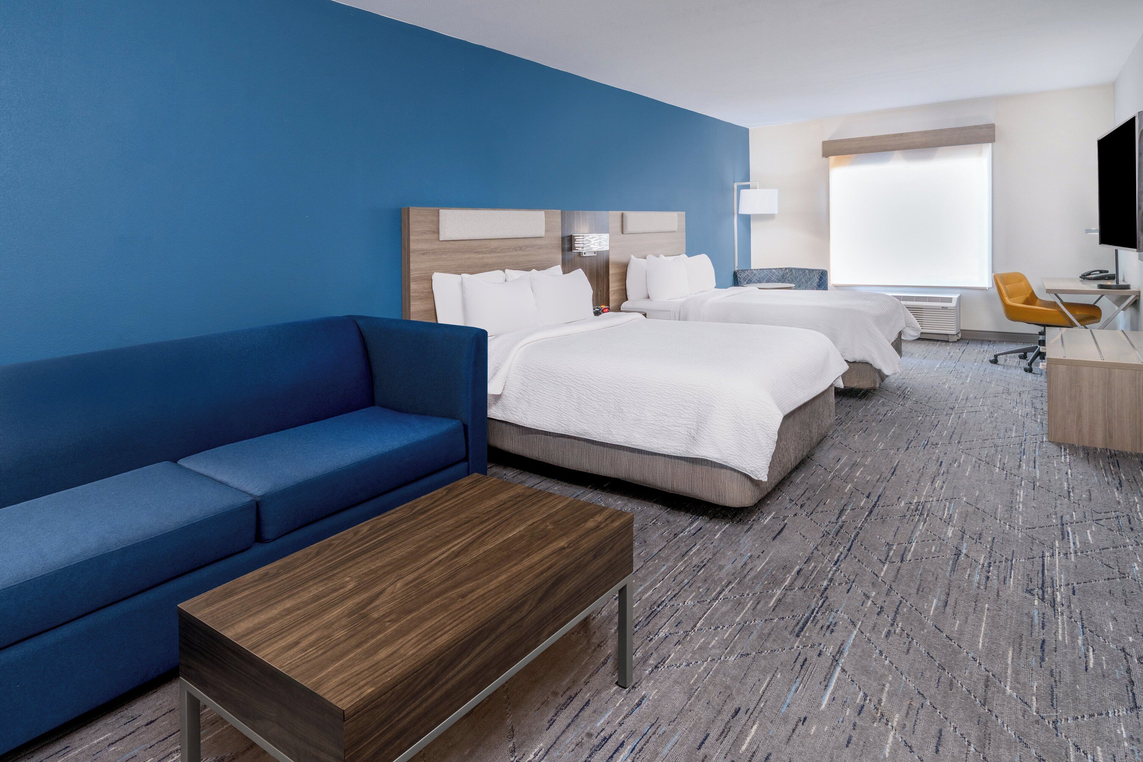 Une chambre d’hôtel avec un canapé bleu, deux lits, une table basse en bois, un bureau avec une chaise et une grande fenêtre.