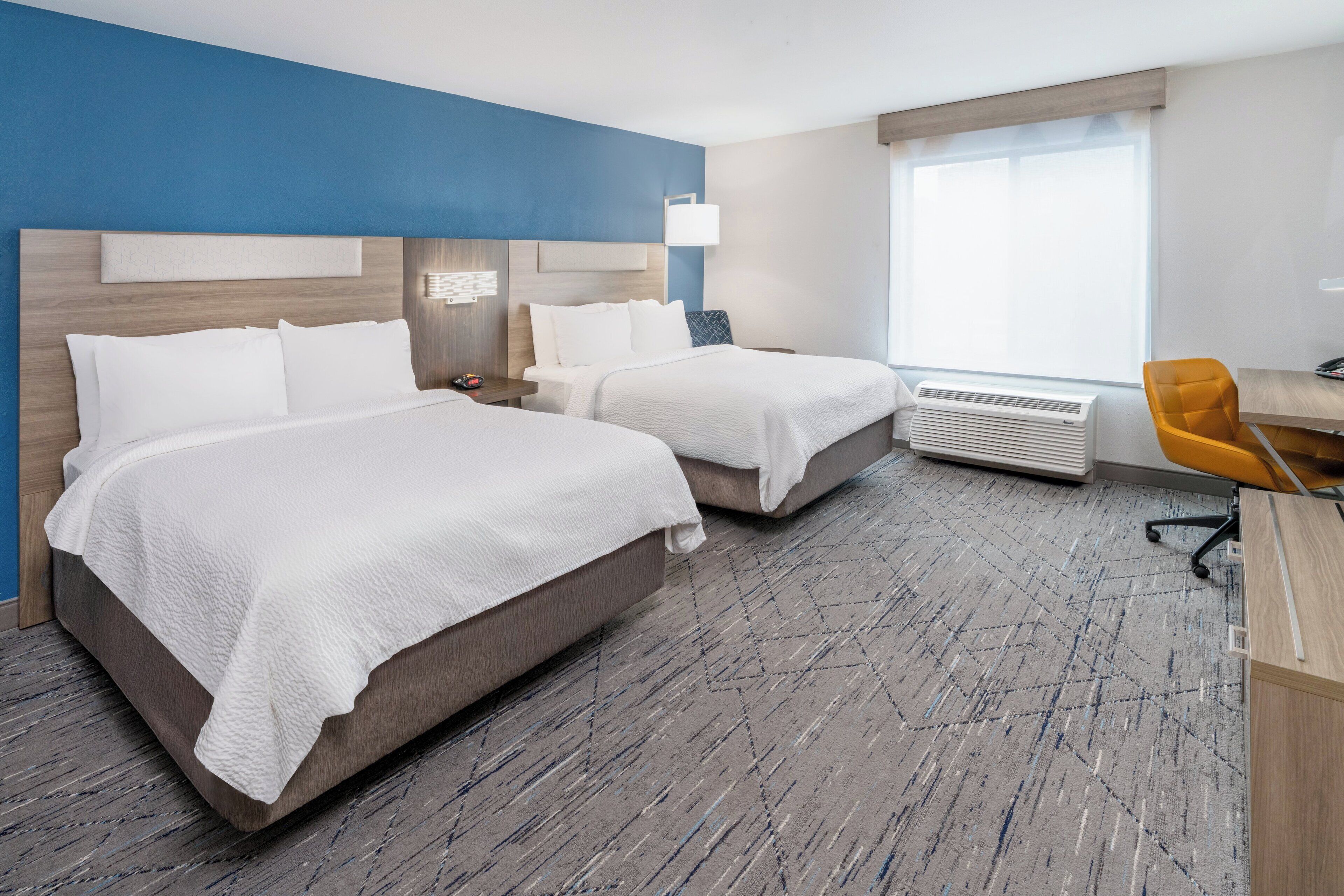 A hotel room with two beds, a desk, an air conditioner, and a window.