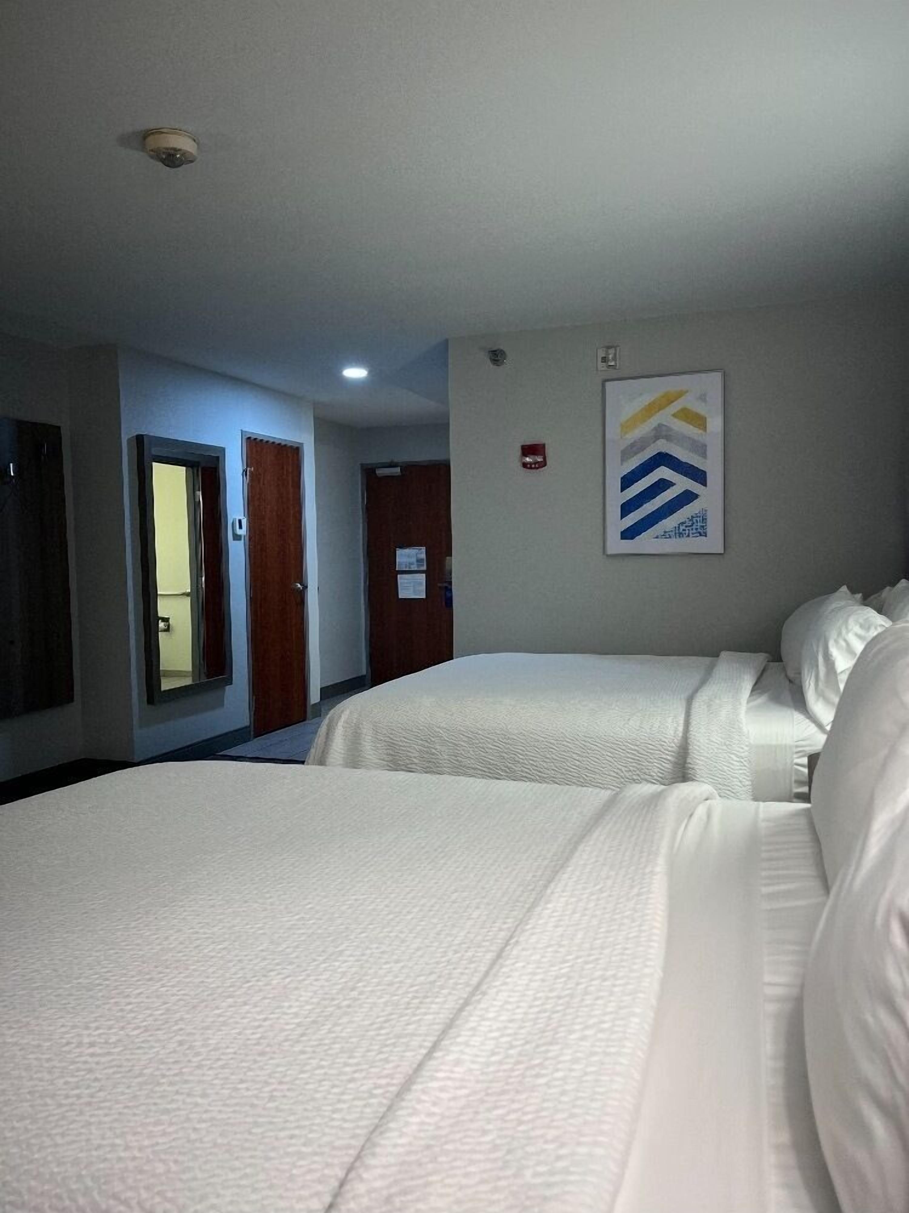 Une chambre d’hôtel avec deux lits, une porte dotée d’un miroir, une œuvre d’art accrochée au mur et un luminaire au plafond.