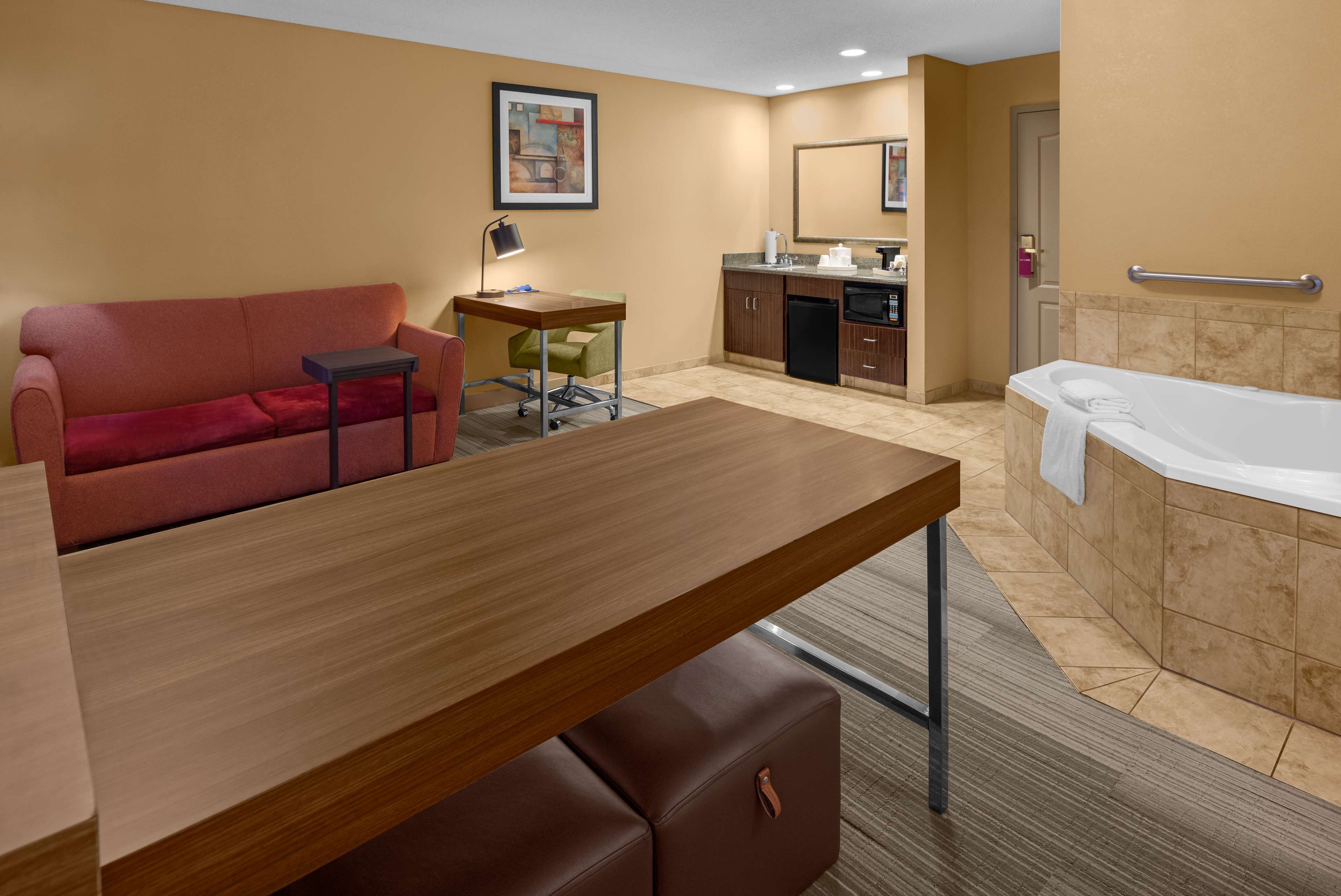 Quarto de hotel com sofá, secretária pequena com luminária, cozinha compacta e banheira.