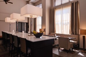 A modern hotel dining area with a long rectangular table, multiple chairs, and a central island with a fruit bowl. There are large floor lamps and a ceiling fan. - Hampton Inn & Suites Banning-Beaumont (Banning)