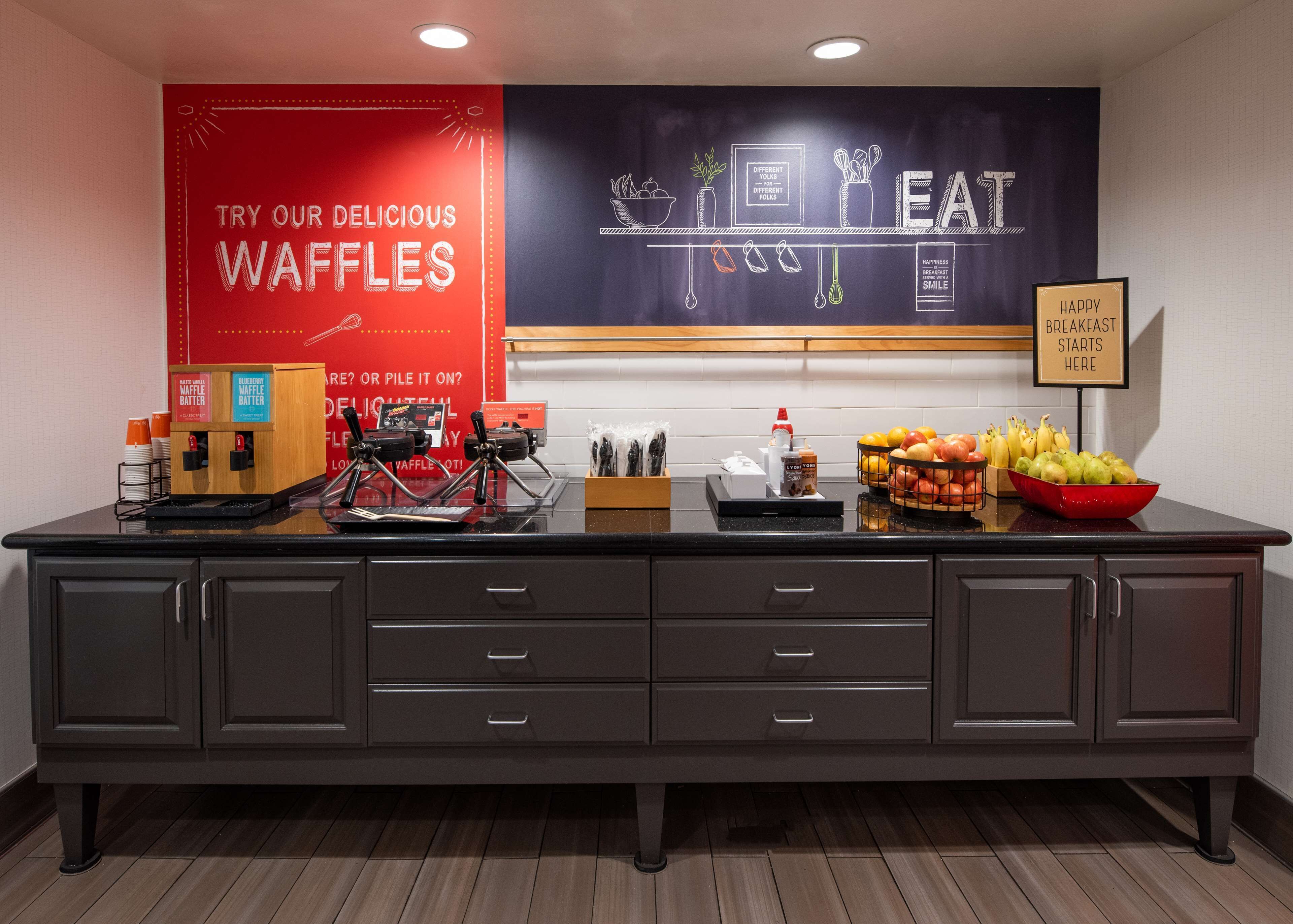 Una barra de buffet con dispensadores de masa para gofres, un bol de fruta y un cartel que indica que el desayuno comienza aquí.