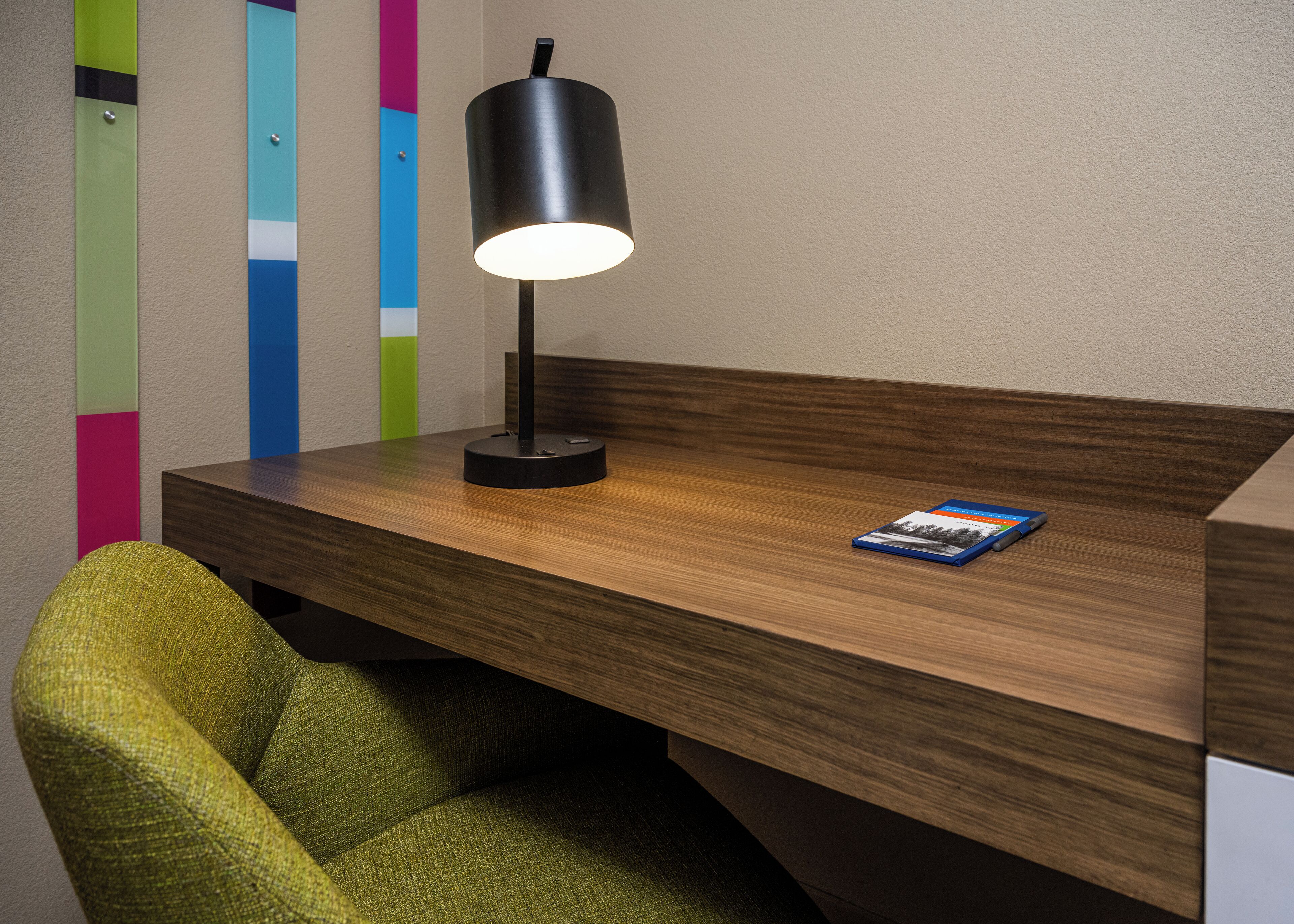 A wooden desk with a black lamp and a book, a green chair, and a wall with colorful vertical stripes.