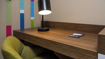 Un escritorio de madera con una lámpara negra y un libro, una silla verde y una pared con rayas verticales coloridas.