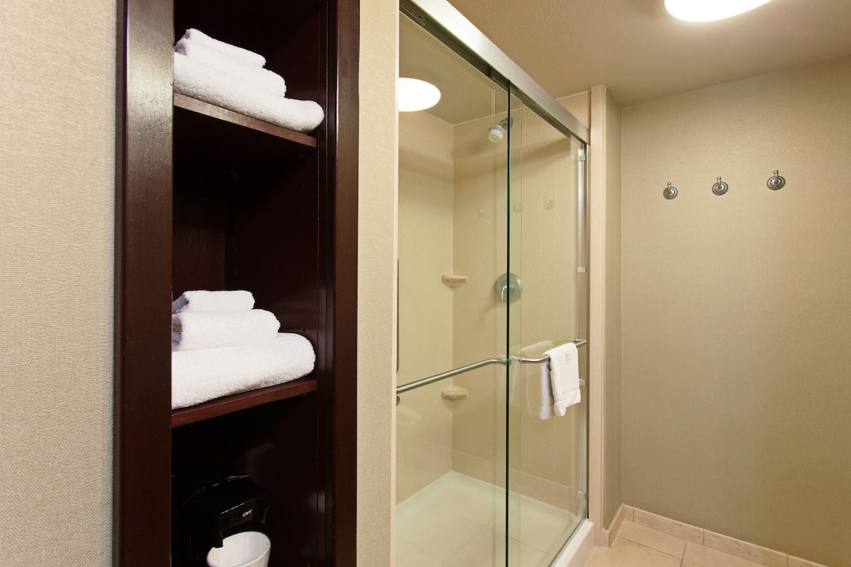 A hotel bathroom with a glass-enclosed shower, a wooden shelf with folded towels, and a beige wall.