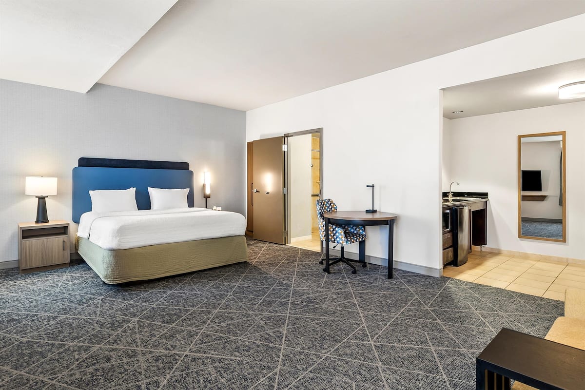 A hotel room with a bed, a desk, a chair, a small table, a mirror, and a kitchenette.